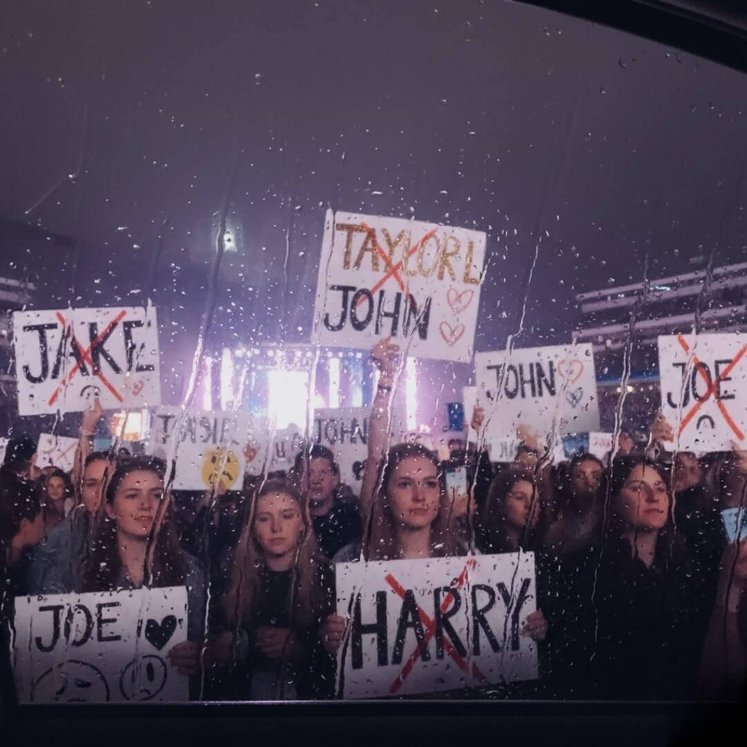 Rain-streaked window view of a concert crowd holding up signs with ex-boyfriend names (like John, Jake, Joe, Harry) crossed out.