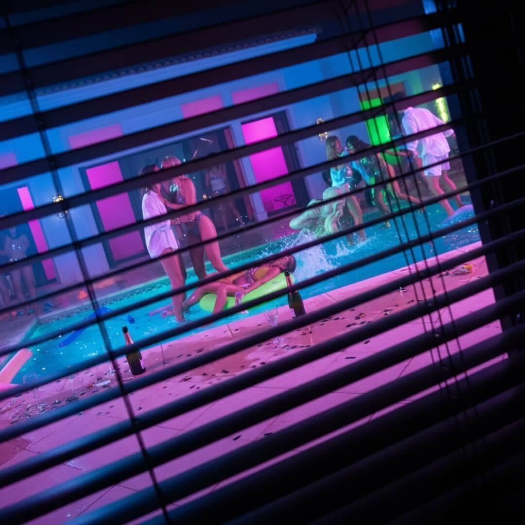 A voyeuristic peek through Venetian blinds at a chaotic villa pool party, lit by neon lights.