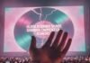 Did Olivia Rodrigo Just Shade Sabrina Carpenter Again? Close-up of a hand with a beaded bracelet unraveling, held up toward a concert screen displaying a cracked vinyl record graphic with the text "OLIVIA RODRIGO SHADE SABRINA CARPENTER RUMORS" in a pink and teal light above a crowd.