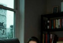 The Influencer Fashion Feud That’s Spilling All Over TikTok A cozy late-night scene from a rainy Seattle apartment: viewed from a low, tilted angle over a steaming red coffee mug on a dark couch, a woman with wide eyes peers at the glowing screen of her open laptop amid bookshelves and rain-streaked windows.